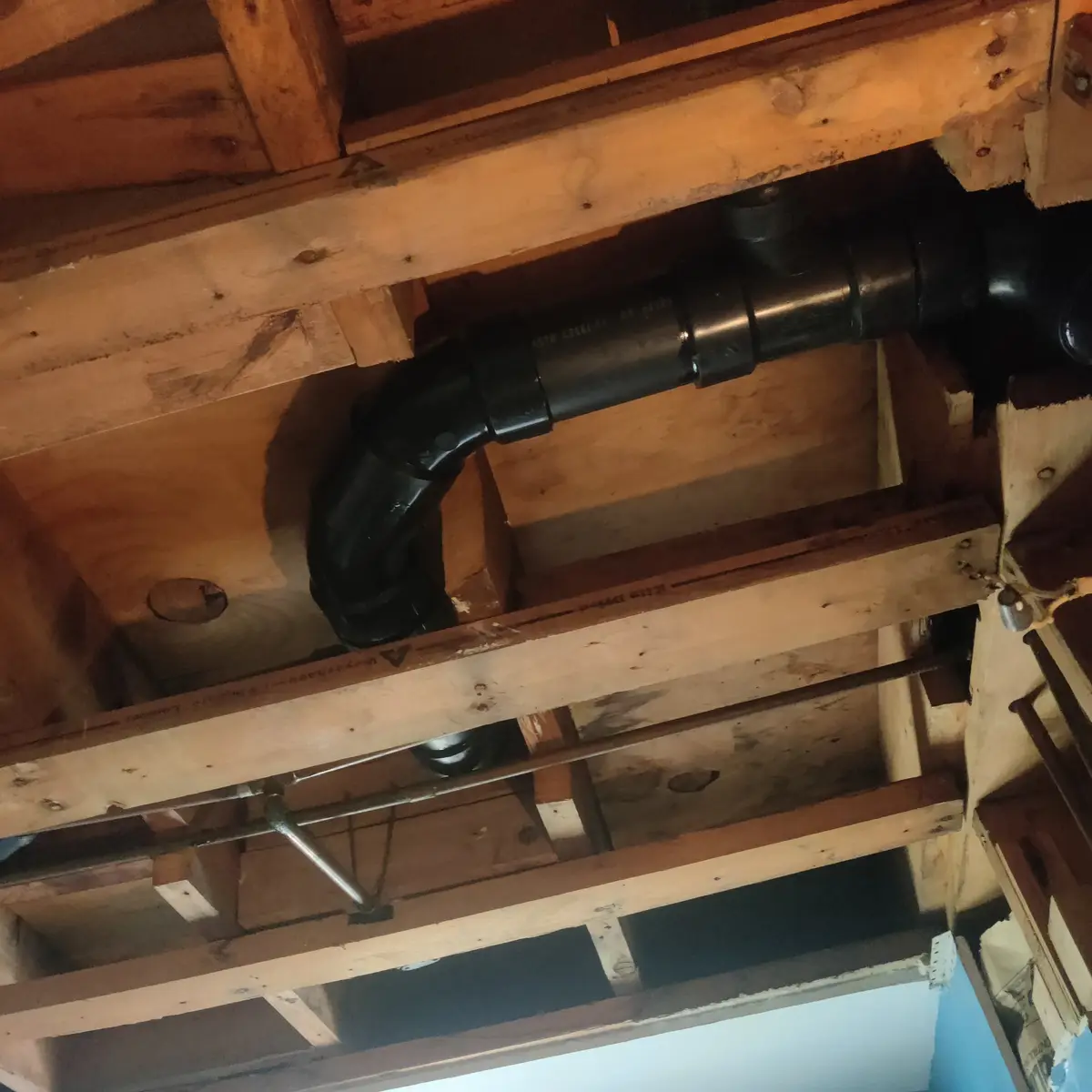 A black pipe runs horizontally through exposed wooden ceiling joists in an attic or crawlspace. The image is taken from…