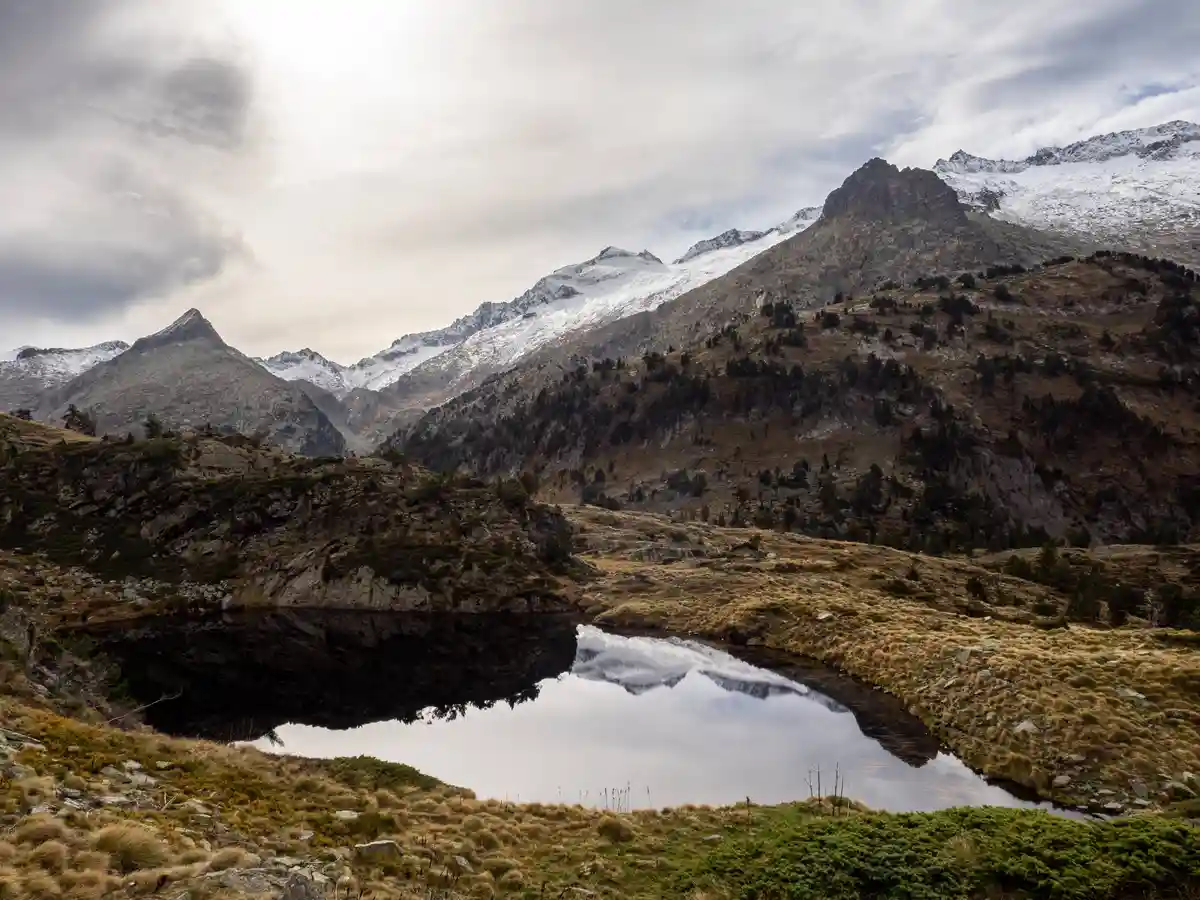 A serene mountain landscape with snow-capped peaks reflected in a calm alpine lake surrounded by rocky terrain and sparse…
