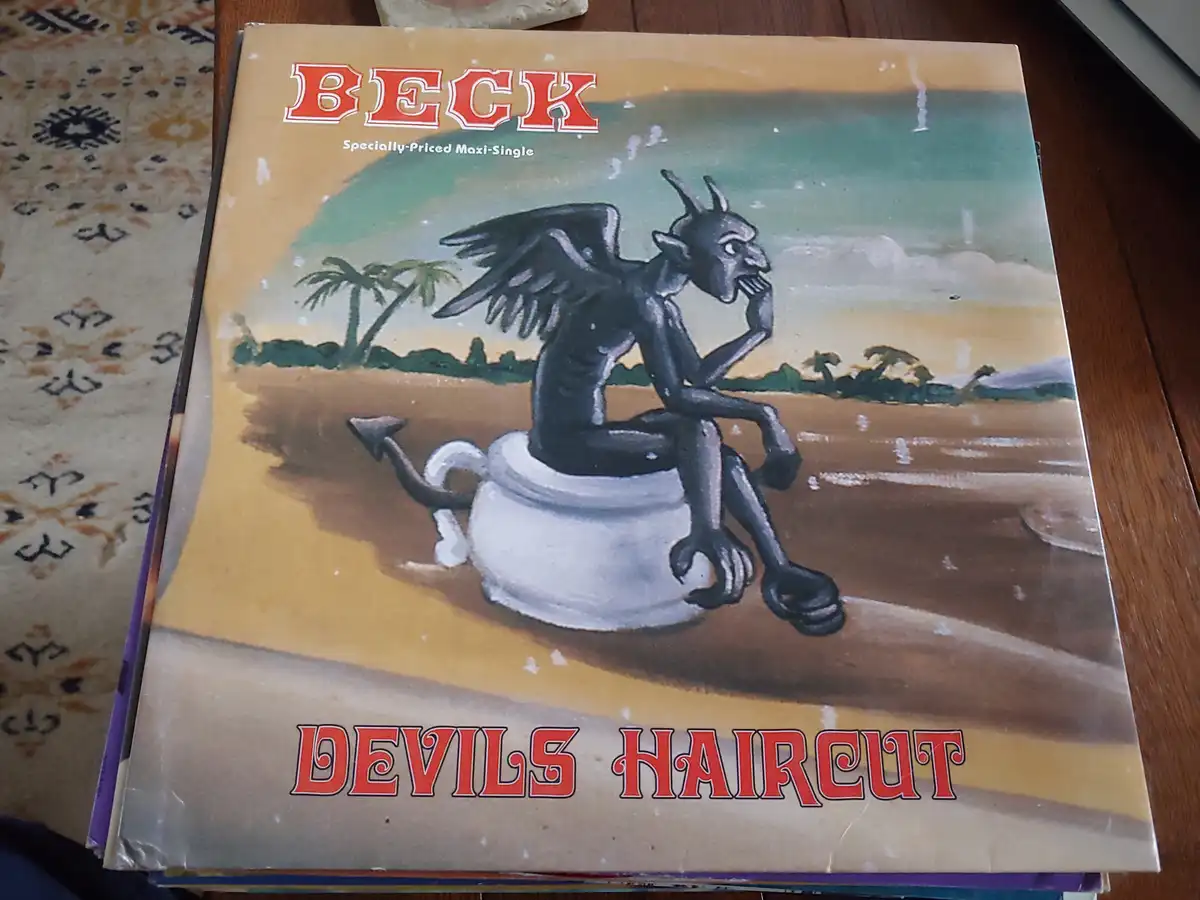 The image shows the cover of Beck's 1994 maxi-single 'Devil's Haircut,' featuring a black-winged, horned devil sitting on a…