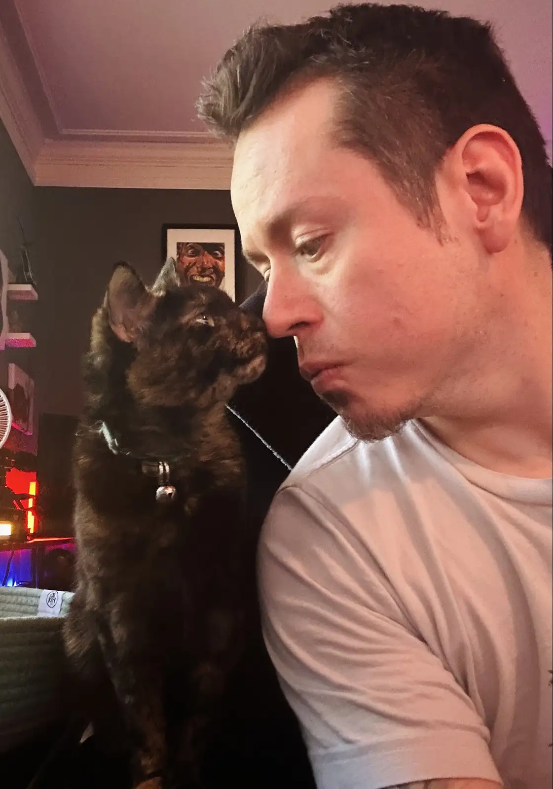 A close-up photo shows a dark tortoiseshell cat standing upright and pressing its nose against the cheek of a man in a white…