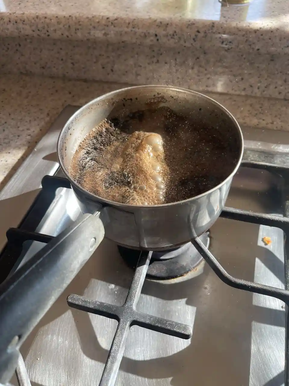A metal saucepan sits on a gas stove burner, containing a dark, bubbling liquid that appears burnt and charred, with a…