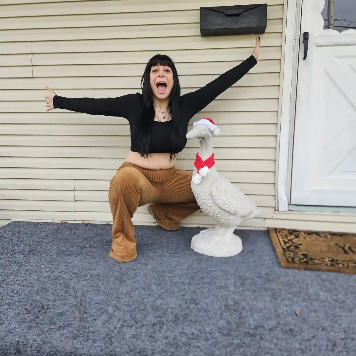 A woman with black hair and bangs, wearing a black crop top and brown corduroy pants, is crouching excitedly on a porch with…