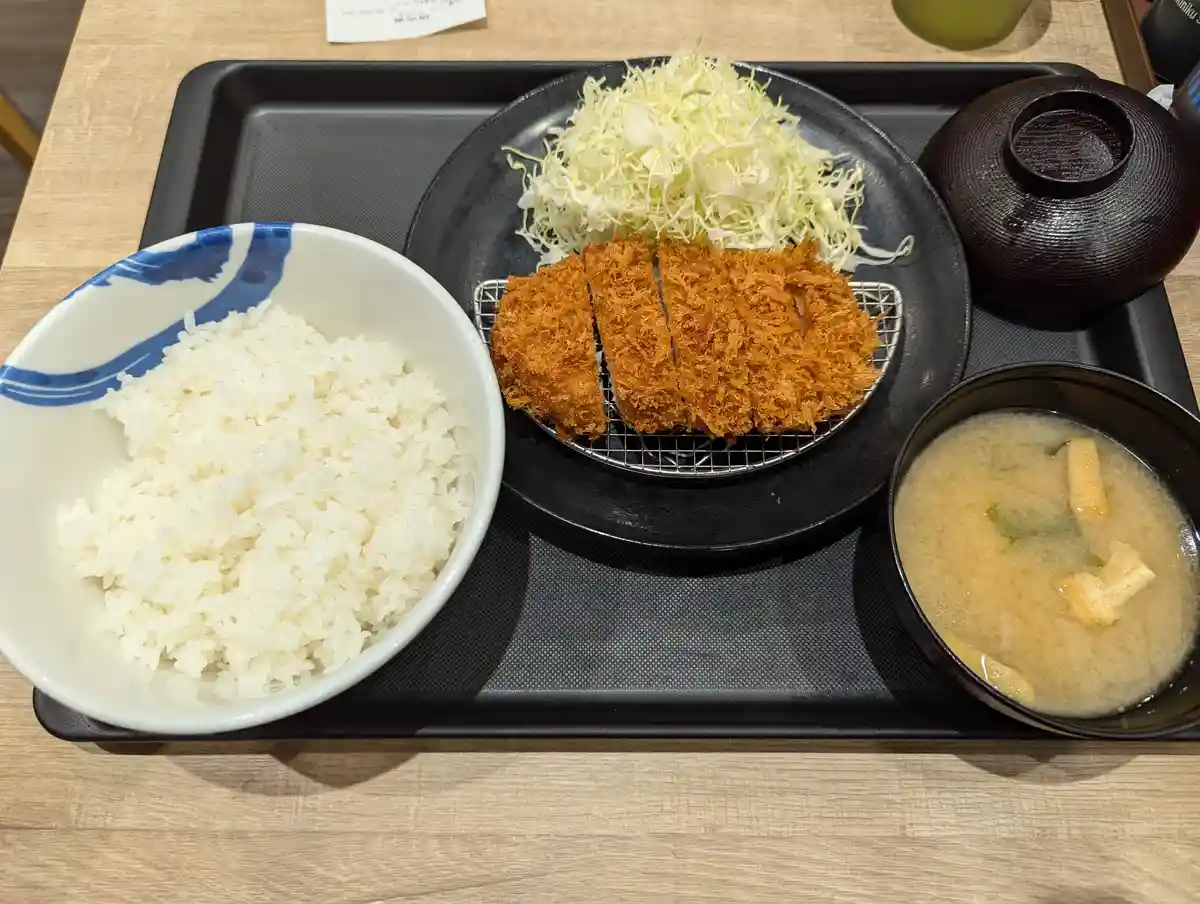 A Japanese meal tray contains a bowl of white rice, a plate of sliced breaded katsu with shredded cabbage, and a bowl of…