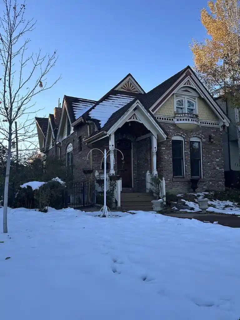 A Victorian-style brick house with snow on its roof and yard, featuring a white porch and bare trees under a clear blue sky…