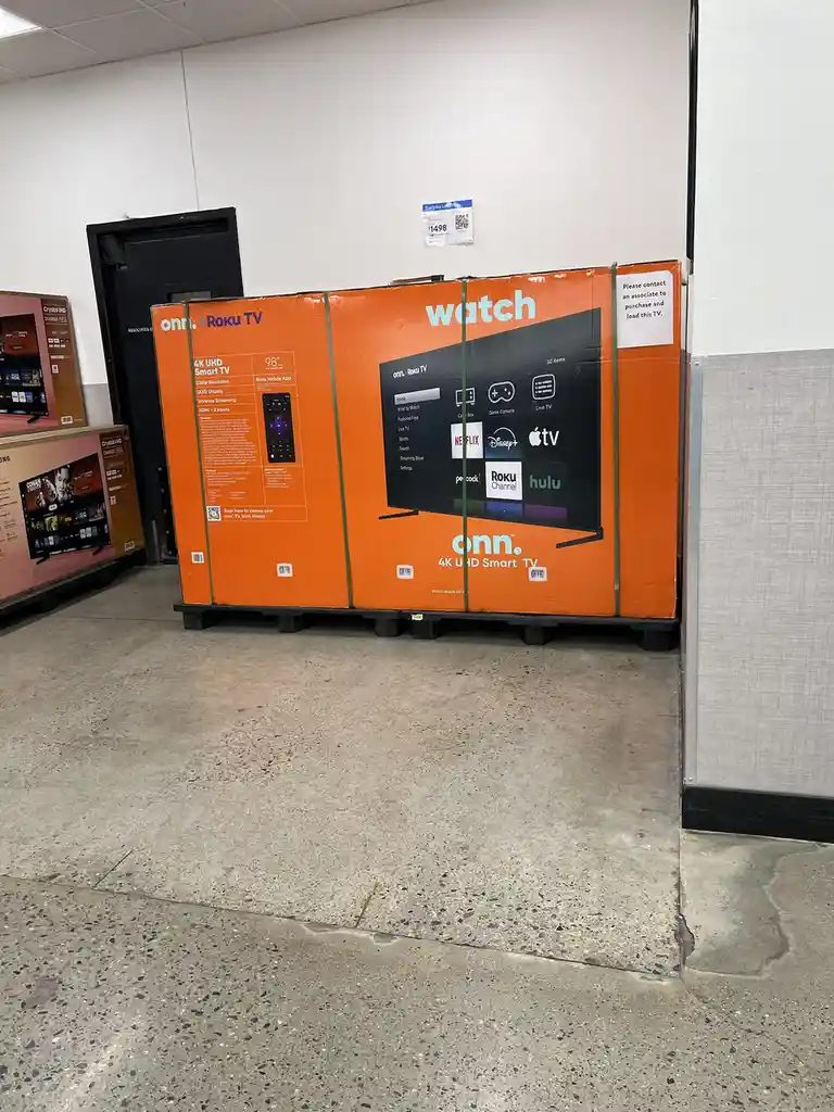 A large orange box for an onn. Roku TV sits on a pallet in a store aisle, with the word 'watch' prominently displayed in…