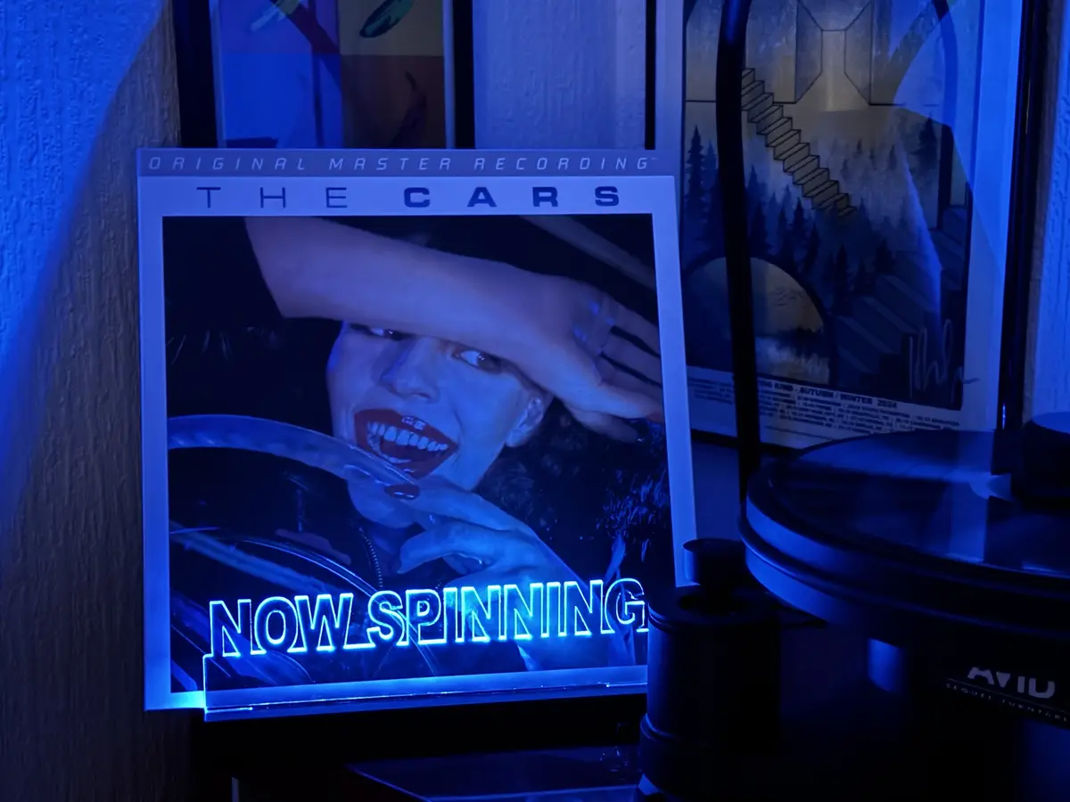 A vinyl record player with a glowing blue 'NOW SPINNING' display is shown next to a framed album cover for 'The Cars'. The…