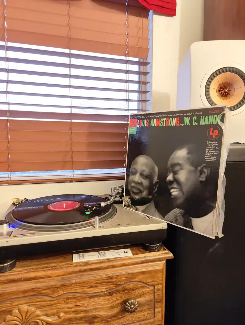 A vintage Technics turntable sits on a wooden dresser with a vinyl record spinning, next to the album cover for 'Louis…