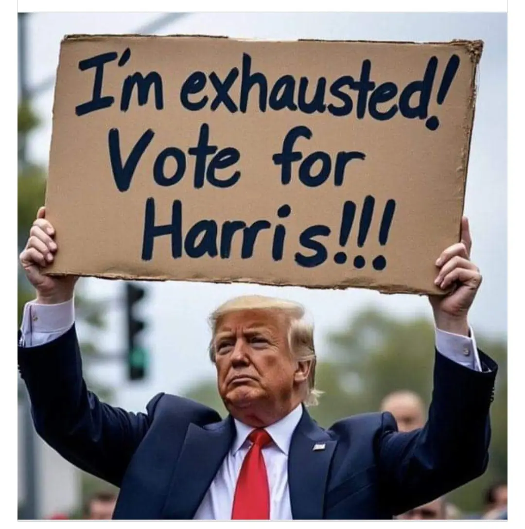 A man in a dark suit and red tie holds up a cardboard sign with black marker text that reads 'I'm exhausted! Vote for…