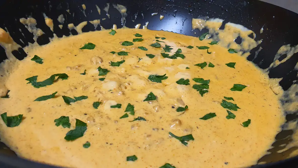 A close-up photo shows a thick, creamy orange-yellow sauce in a black pan, speckled with red spices and garnished with…