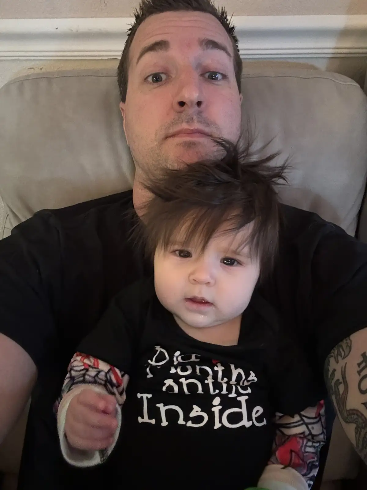 A man with a surprised expression lies on a couch holding a baby who is wearing a black shirt with the text 'Did 9 Months…