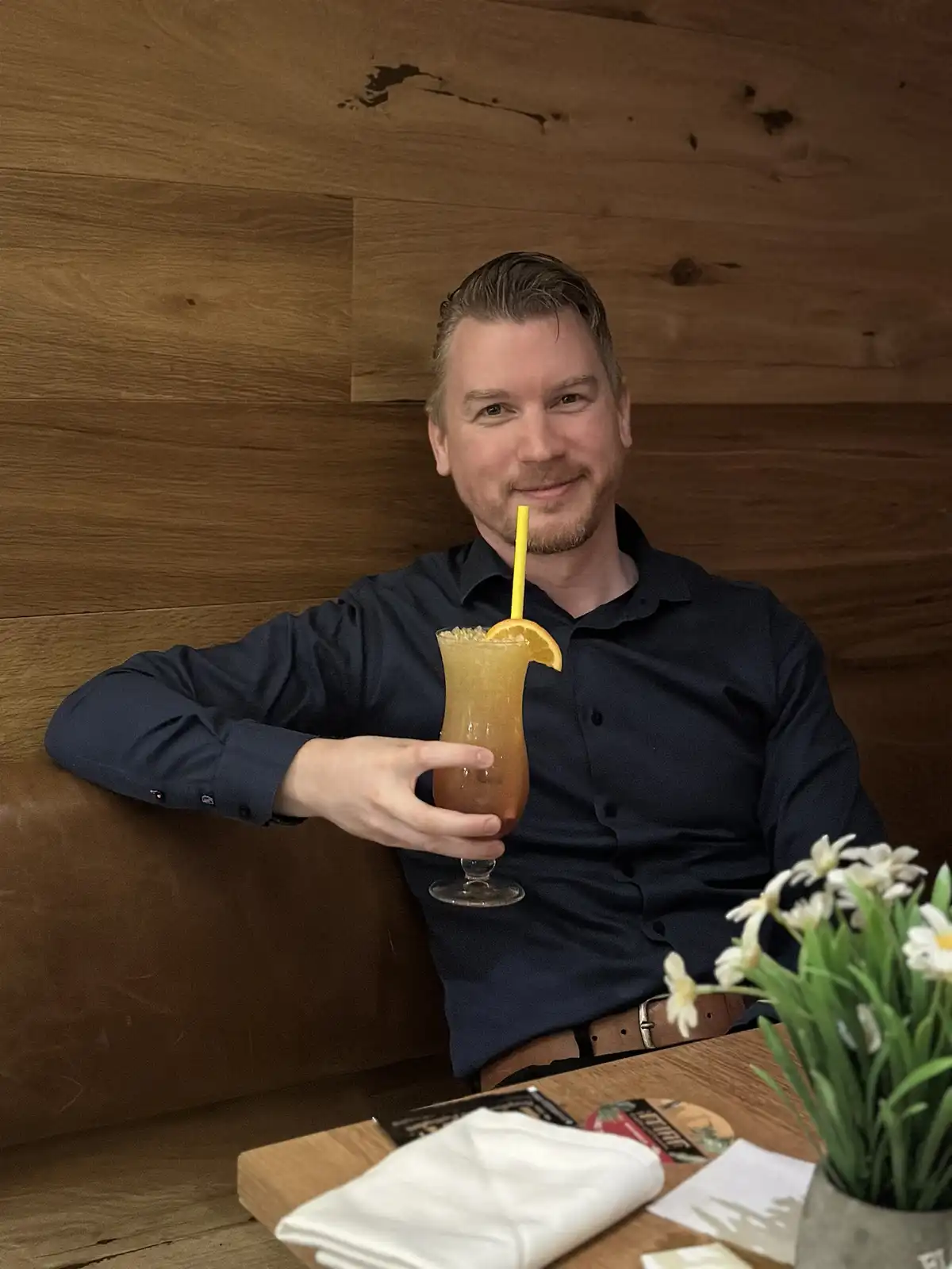 A man in a dark blue button-down shirt sits in a booth with a wooden wall behind him, holding a tall glass of orange-colored…