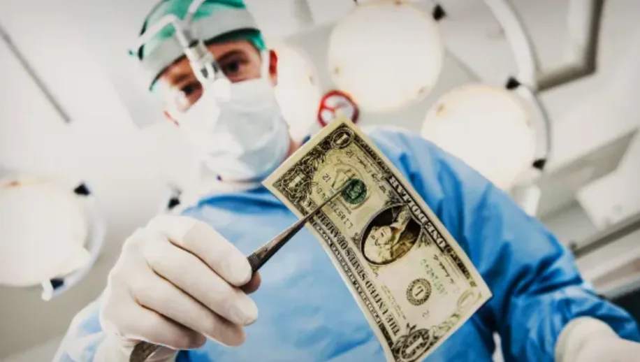 A surgeon in blue scrubs, surgical cap, and face mask holds a one-dollar bill with tweezers in an operating room. The…