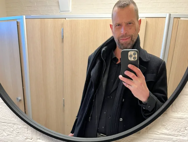 A man in a black coat takes a selfie in a round mirror inside a public restroom, with wooden stall doors visible behind him…