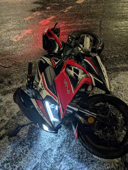 A red, black, and white Lexmoto motorcycle lies on its side on a wet asphalt road at night, its headlight still on and…