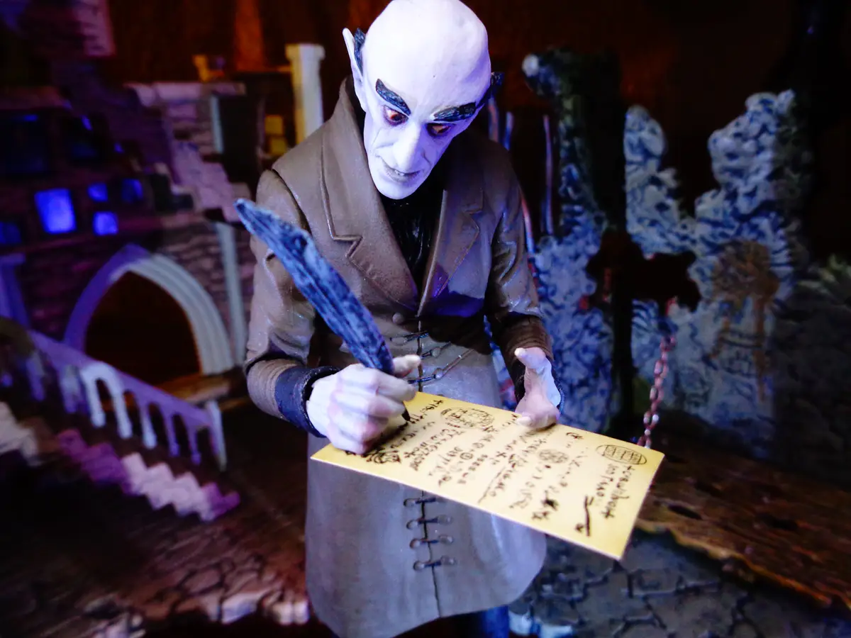 A pale, bald, demonic-looking figurine in a brown coat is writing on a yellow envelope with a quill pen. The background is a…