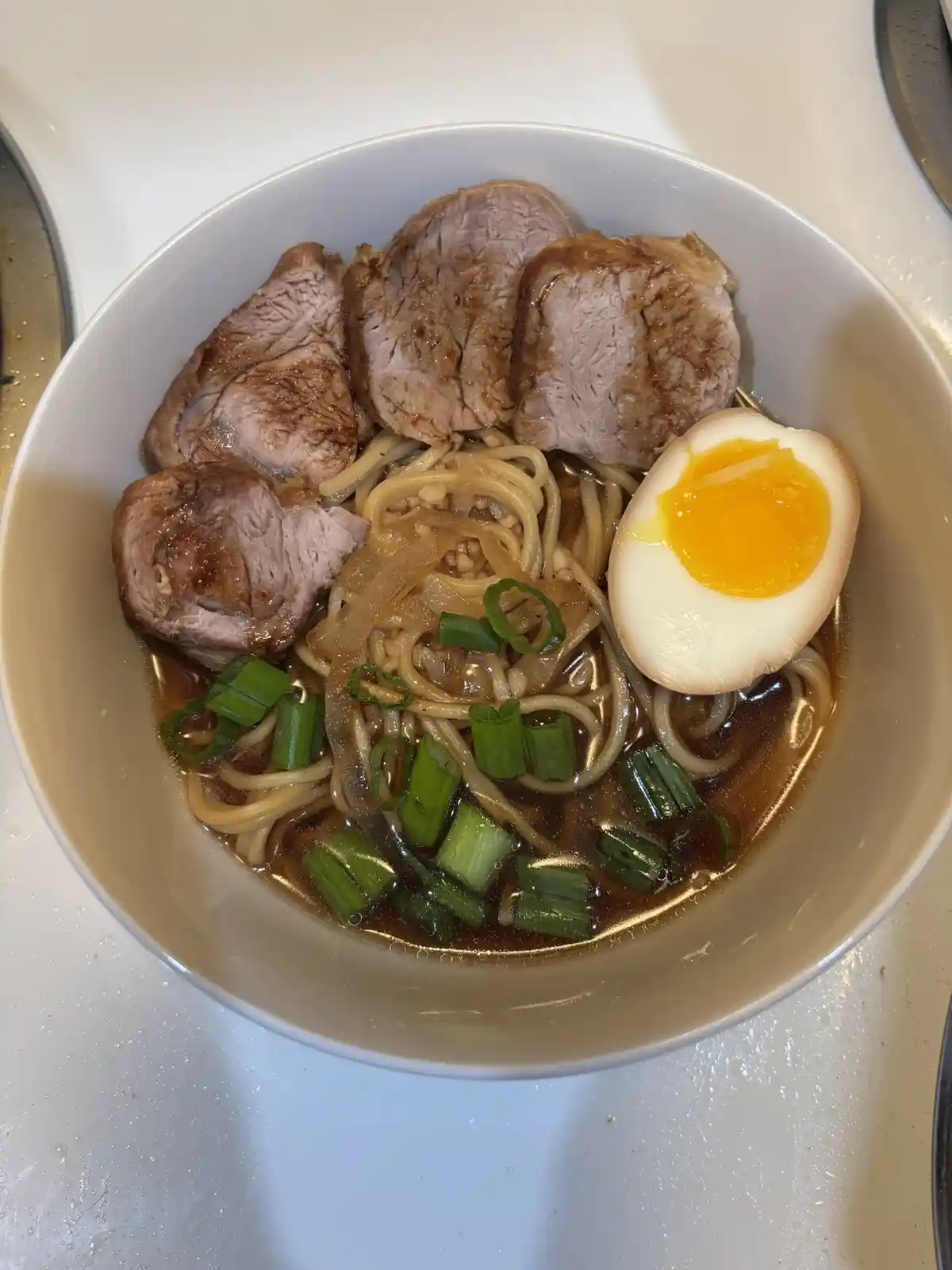 A top-down photo of a white bowl filled with ramen noodles, sliced pork, a soft-boiled egg cut in half, and chopped green…