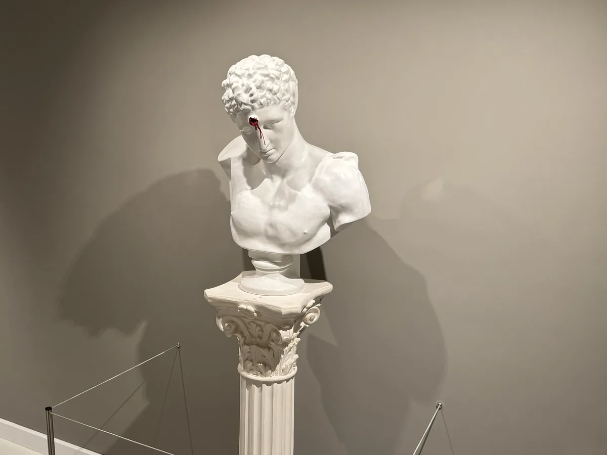 A white marble bust of a classical male figure, displayed on an ornate pedestal in a museum setting, has a single red tear…