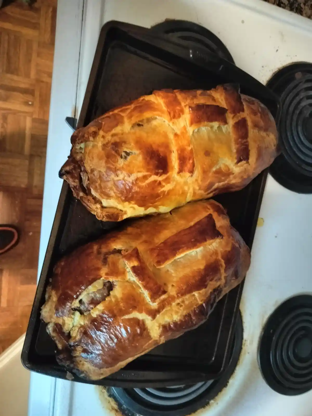 Two golden-brown, flaky pastry-wrapped meatloaves sit on a black baking tray placed on a white electric stovetop. The top…