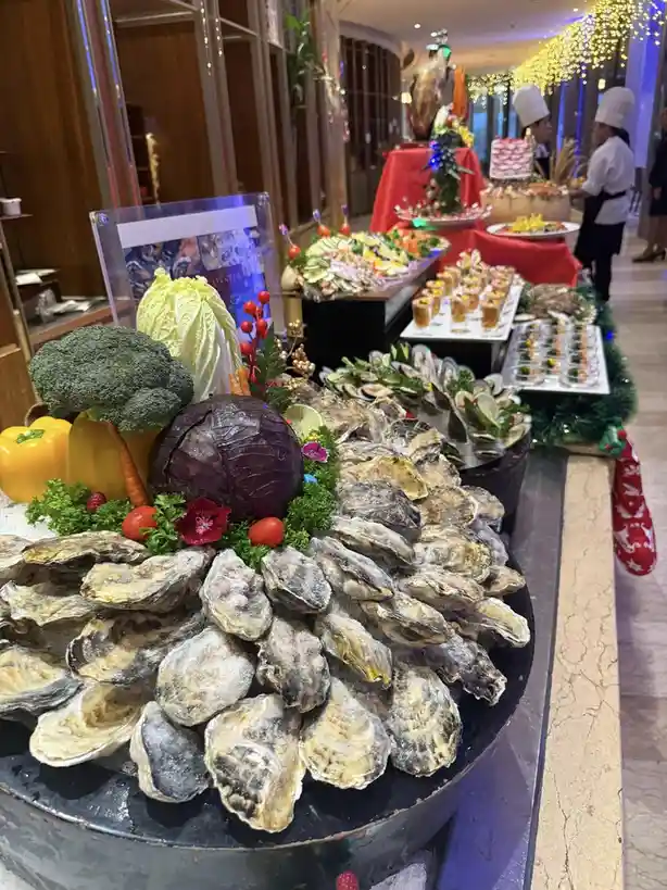 A lavish buffet display features a large black bowl overflowing with raw oysters on the half shell, surrounded by decorative…