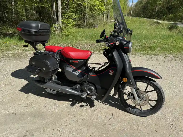 A black Honda Super Cub motorcycle with red seat and accents is parked on a gravel road beside green grass and trees. It has…