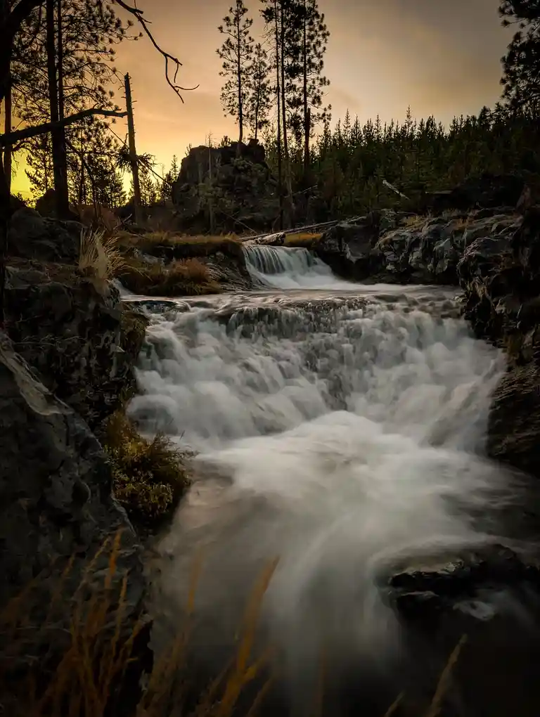 A long-exposure photograph of a cascading mountain stream at sunset, with smooth, blurred water flowing over dark rocks…