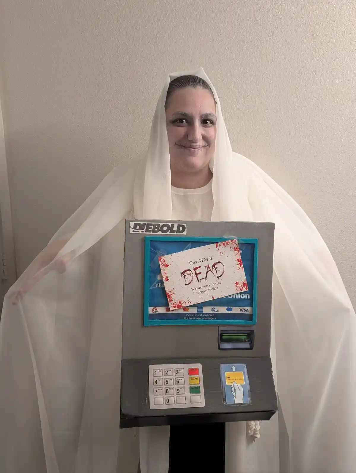 A person dressed in a white, flowing robe resembling a nun is holding a cardboard cutout of a Diebold ATM. The ATM screen…