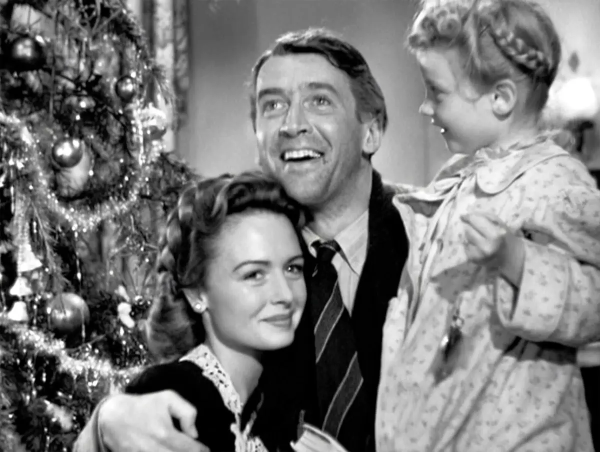A black-and-white photo from a classic Christmas movie shows a smiling man holding a young girl, with a woman beside them…