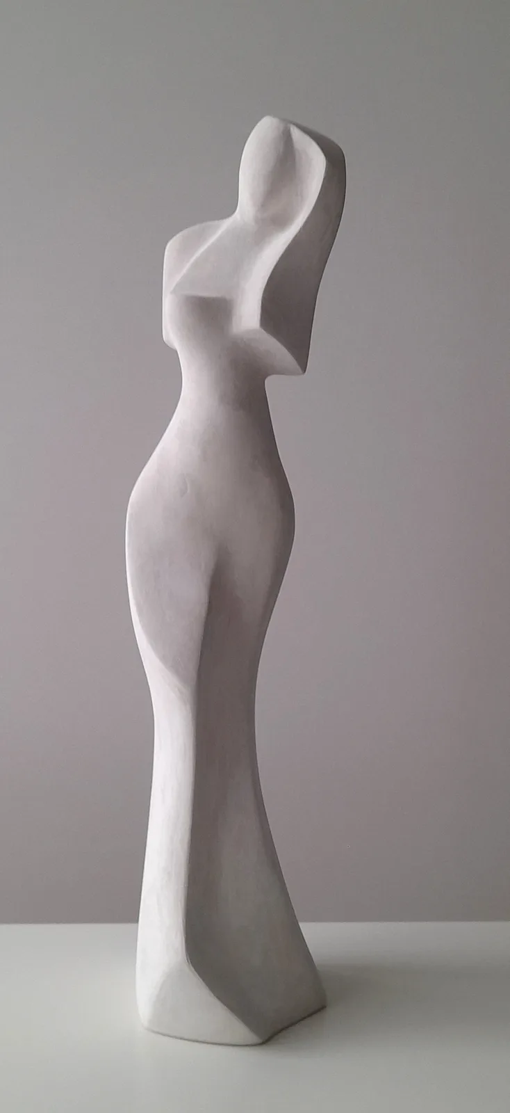A minimalist white abstract sculpture of a human figure viewed from the side, standing on a white surface against a plain…