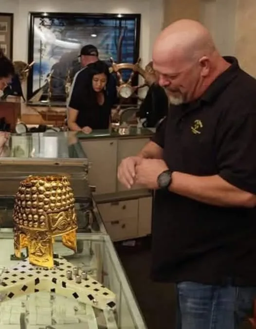 A bald man in a black polo shirt stands in front of a glass display case containing a golden, ornate helmet-like artifact…