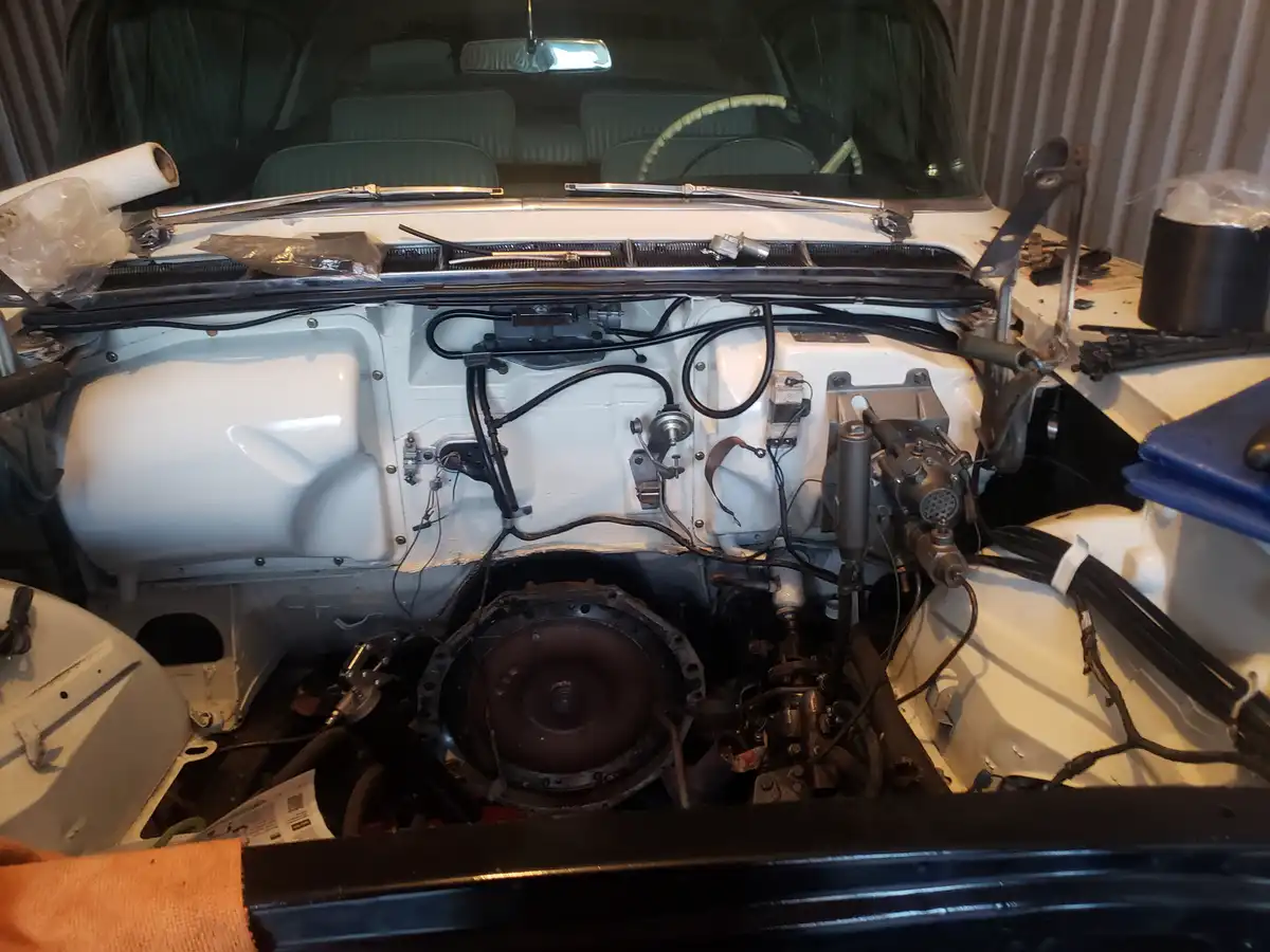 A white vintage car's engine bay is shown with the hood open, revealing exposed wiring, mechanical parts, and a partially…