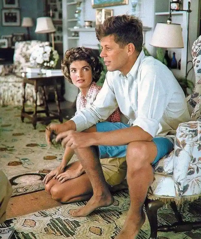 A vintage photo shows John F. Kennedy sitting on a floral-patterned chair with his legs crossed, wearing a white shirt and…