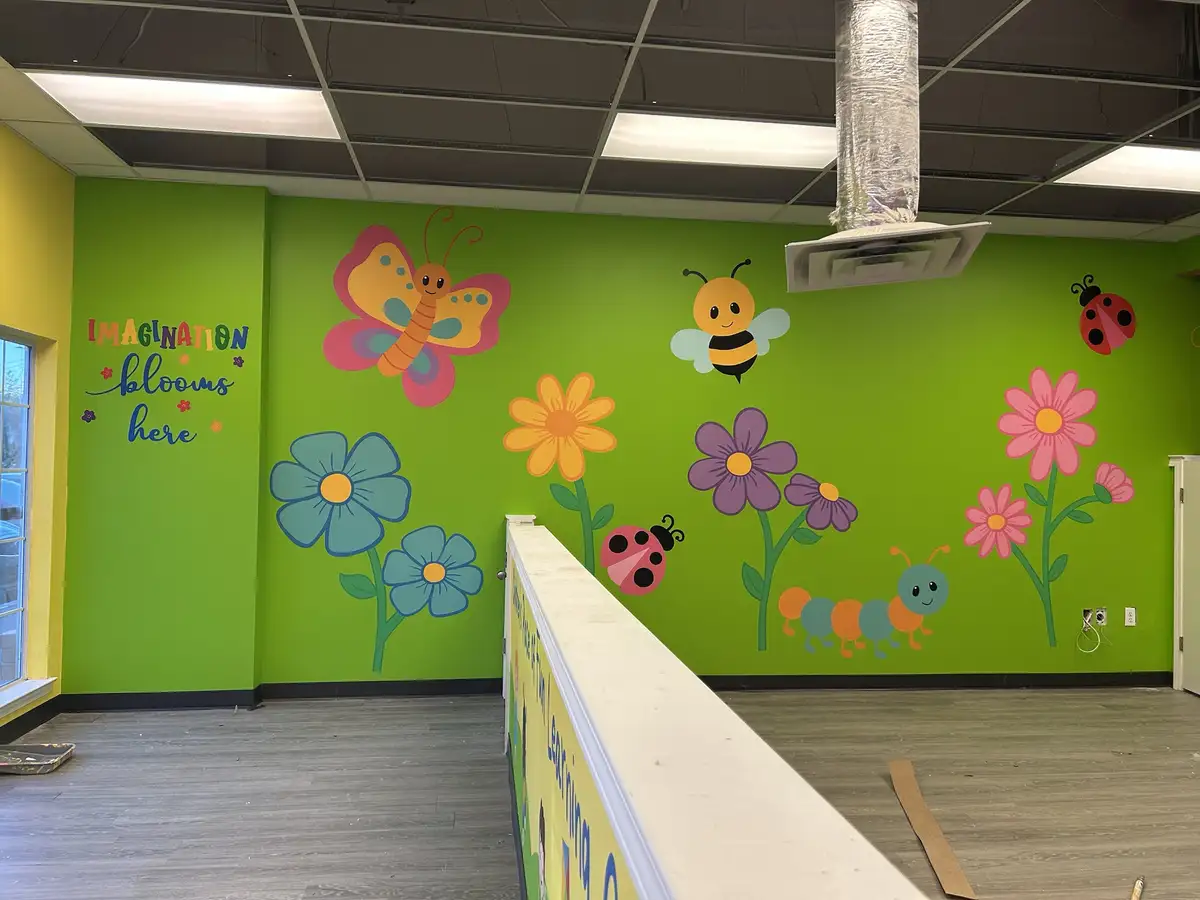 A brightly painted green wall in what appears to be a children's playroom or classroom features cartoon-style murals of a…