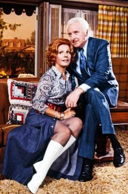 A vintage photo shows a man and woman sitting on a couch in a retro living room. The woman, with red hair and wearing a…