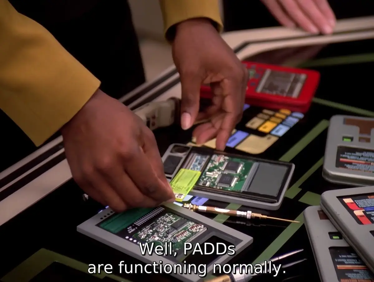 A close-up shot shows a person in a yellow uniform handling disassembled PADD devices on a console, with overlaid text…