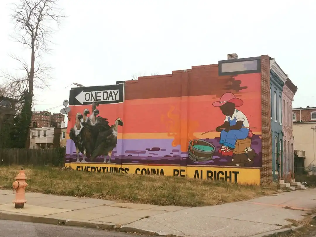 A colorful mural on a brick building shows a child fishing with a bucket while a flock of vultures stands nearby. Above the…