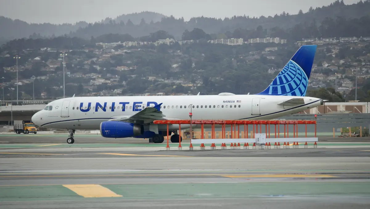 A United Airlines passenger jet is parked on an airport tarmac with a red jet bridge partially attached to its door. The…