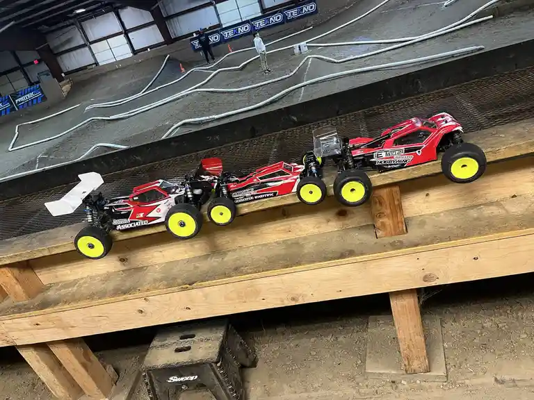 Three red and white remote-controlled monster trucks with bright yellow wheels are lined up on a wooden bench overlooking an…