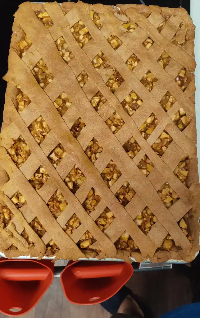 A large rectangular apple pie with a golden-brown lattice crust sits on a baking sheet, its filling visible through the…