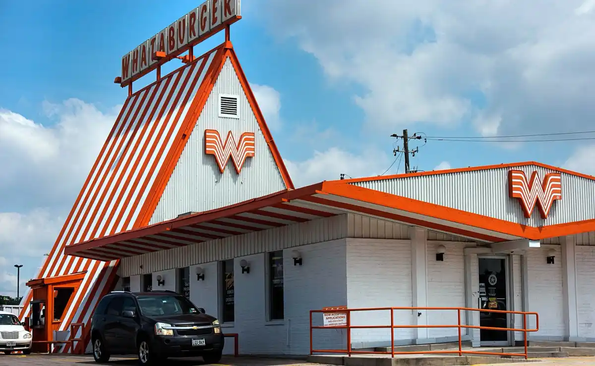 An exterior photo of a Whataburger restaurant with its iconic orange and white striped roof and large 'W' logo, under a…