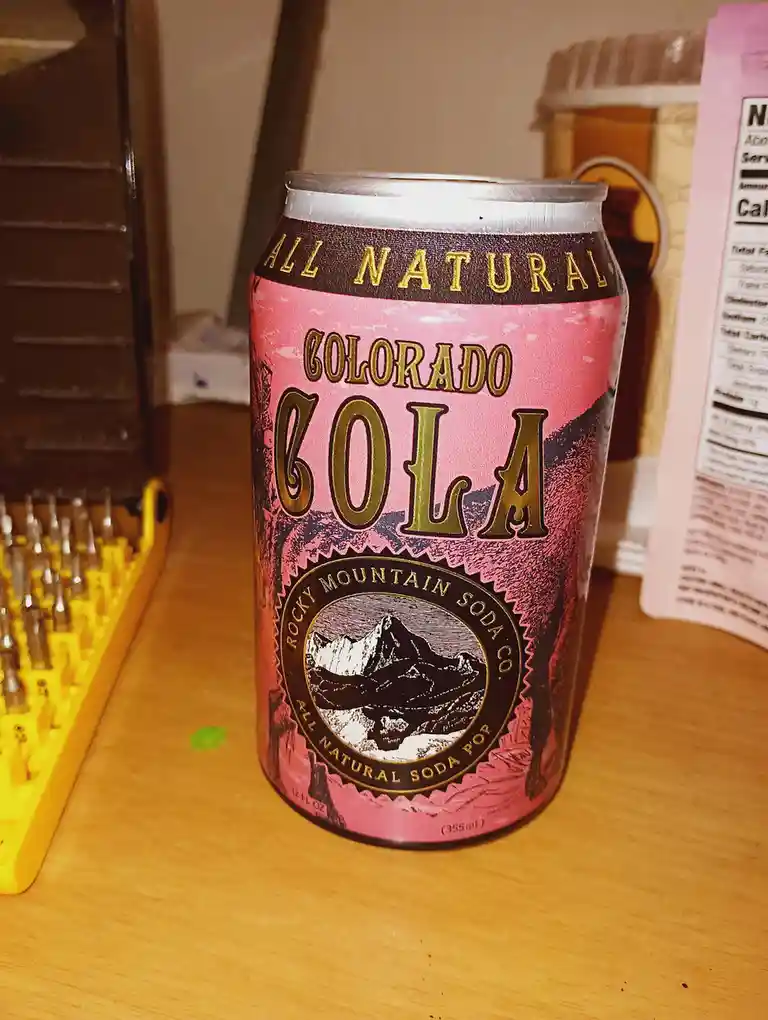 A can of 'Colorado Cola' by Rocky Mountain Soda Co. sits on a wooden surface, featuring a pink label with gold lettering and…