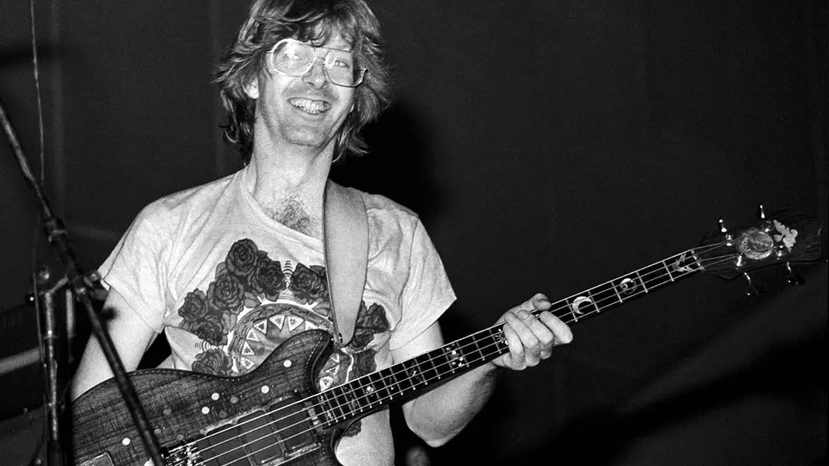 a black and white photo of a smiling man with glasses and shoulder-length hair playing a bass guitar. He wears a t-shirt…