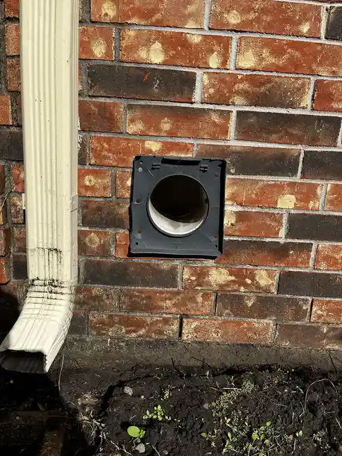 A close-up photo shows a black square vent cover with a circular opening, mounted on a weathered red brick wall next to a…