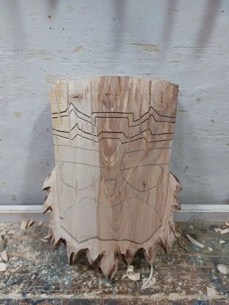 A wooden carving in progress, showing a rough, partially carved face with deep grooves and pencil outlines, sitting on a…