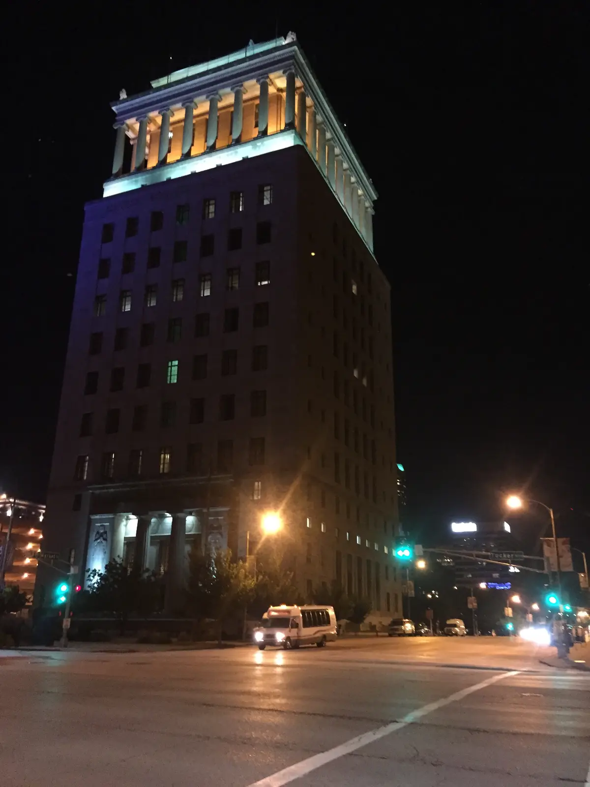 A nighttime photo of a tall, classical-style building with a lit-up top section and a dark lower section, with streetlights…