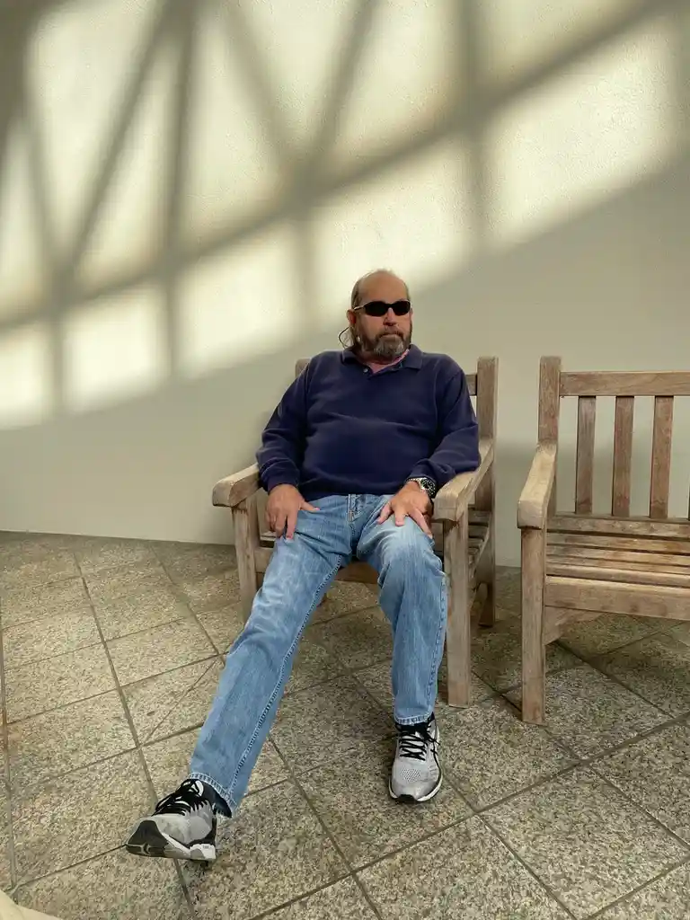 A middle-aged man with a beard and sunglasses sits relaxed on a wooden bench, one leg crossed over the other, wearing a blue…