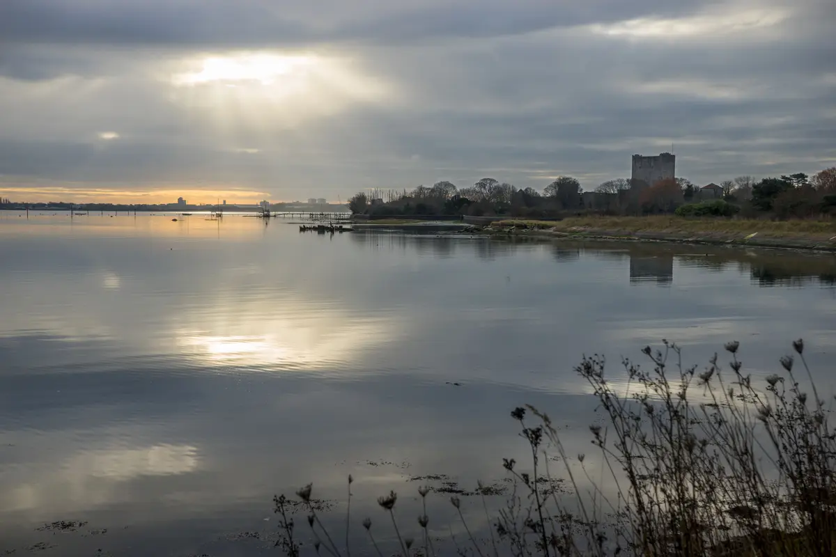 A serene landscape photo shows a calm body of water reflecting a cloudy sky with sunlight breaking through. On the distant…