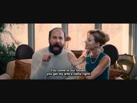 A man with a beard and a woman in a blue dress are in a living room. The man is pointing at the woman while she touches his…