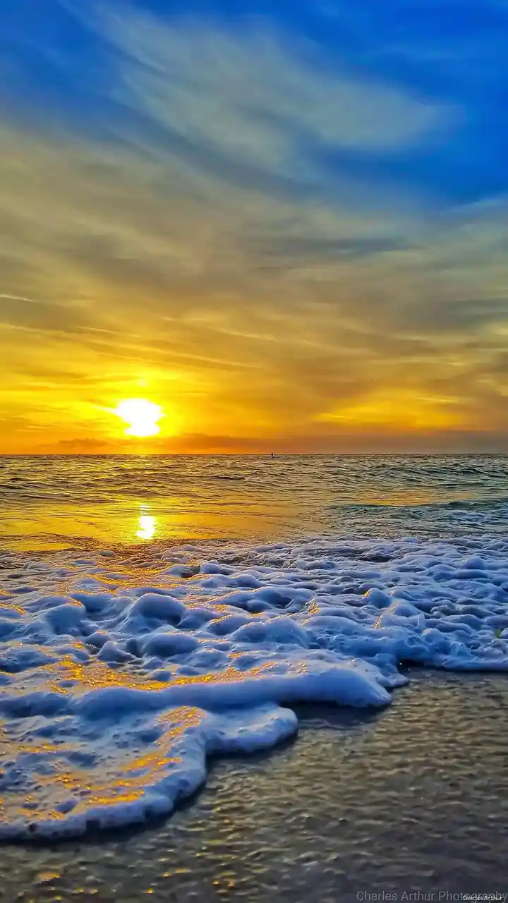 A vibrant beach sunset photo shows golden sunlight reflecting on ocean waves and foamy surf, with a blue sky and wispy…