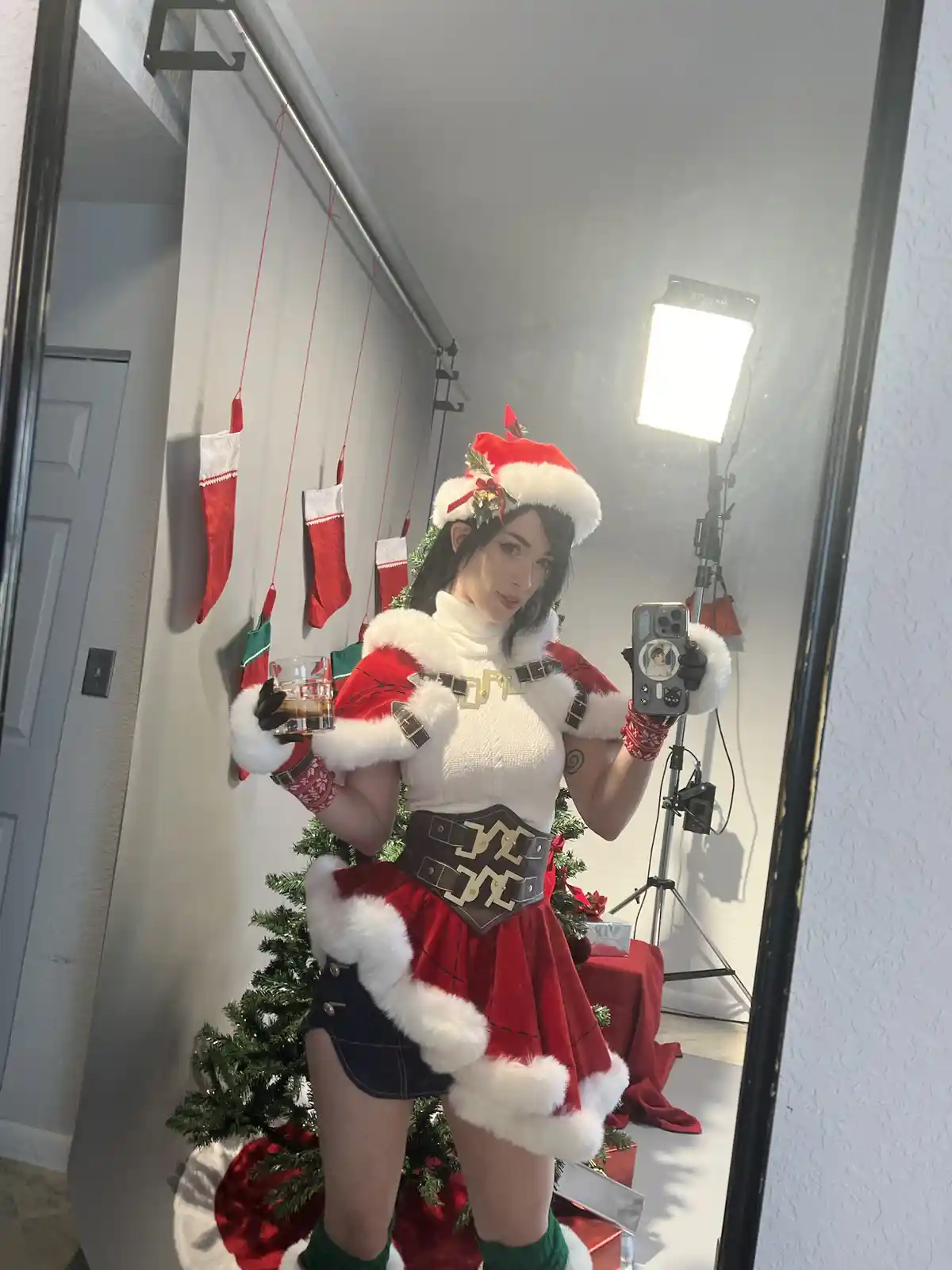 A person in a stylized Santa costume with a white turtleneck, red skirt, and fur trim takes a mirror selfie while holding a…