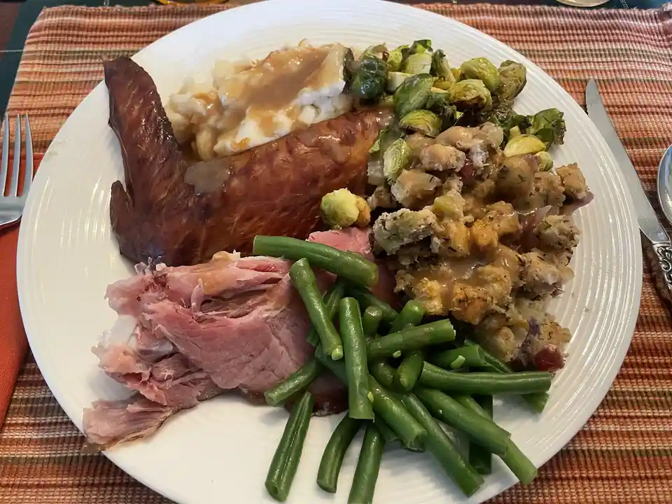 A white plate holds a large, roasted ham with a dark, crispy skin, alongside mashed potatoes with gravy, roasted brussels…